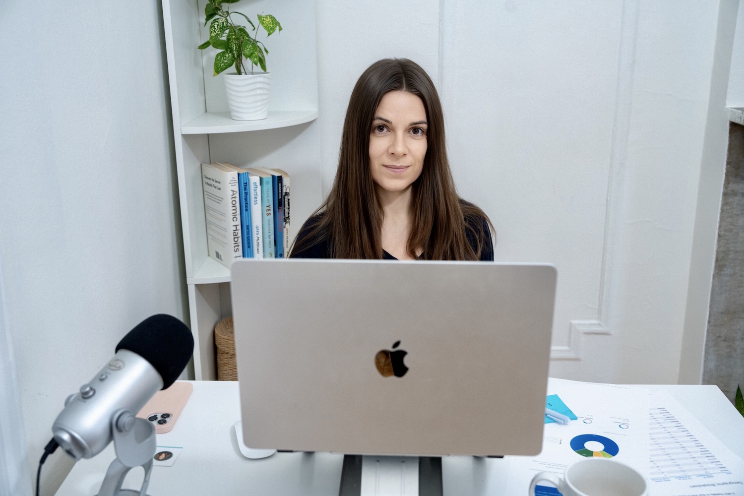 Woman with long brown hair sitting at desk behind laptop with microphone, books, and documents nearby.
