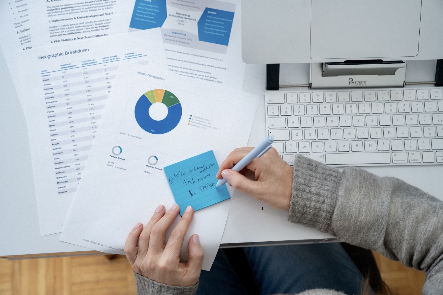Person writing on blue sticky note over business documents with charts near a laptop keyboard.