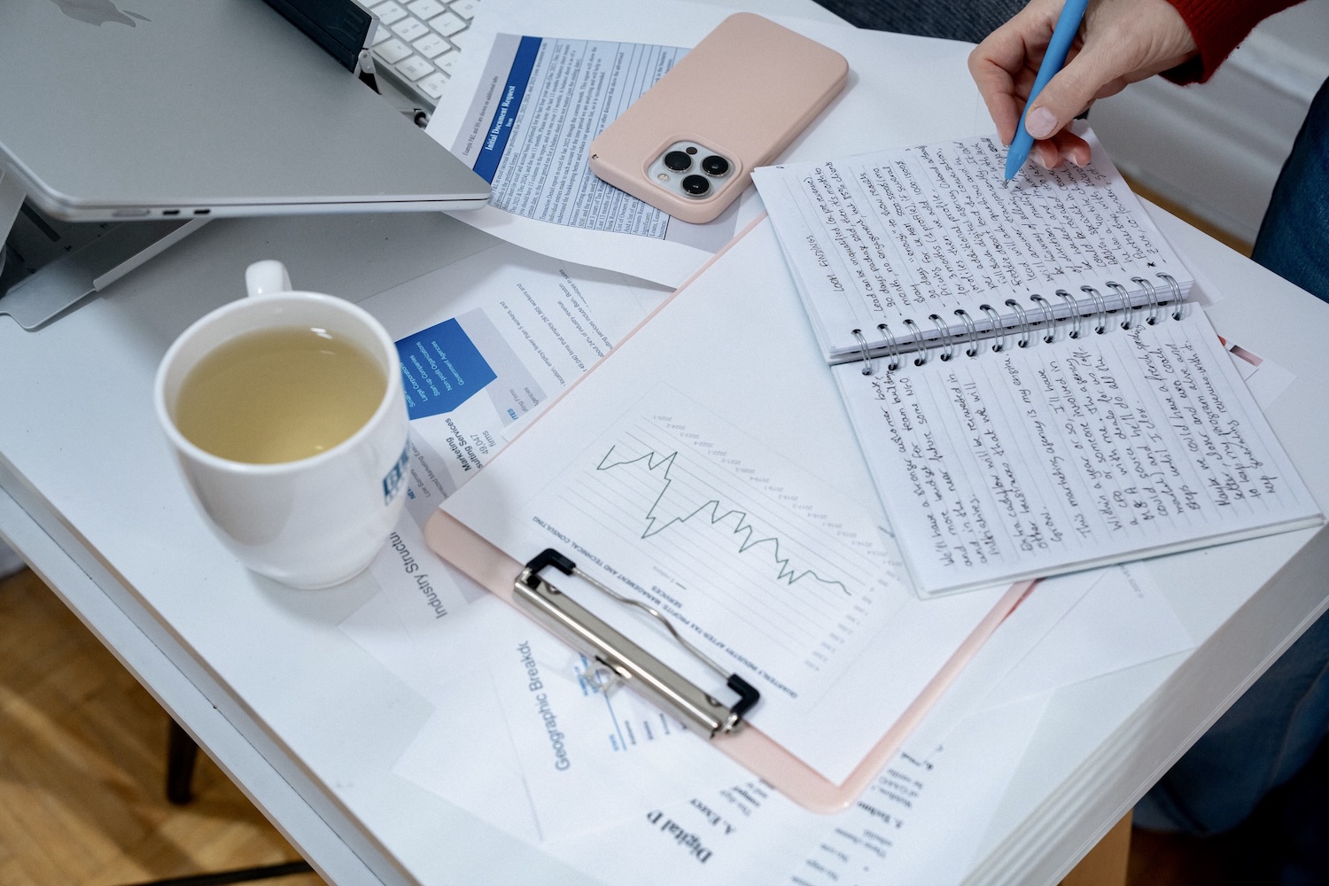Person writing notes with a blue pen on a spiral notebook beside papers, phone, laptop, and a cup.