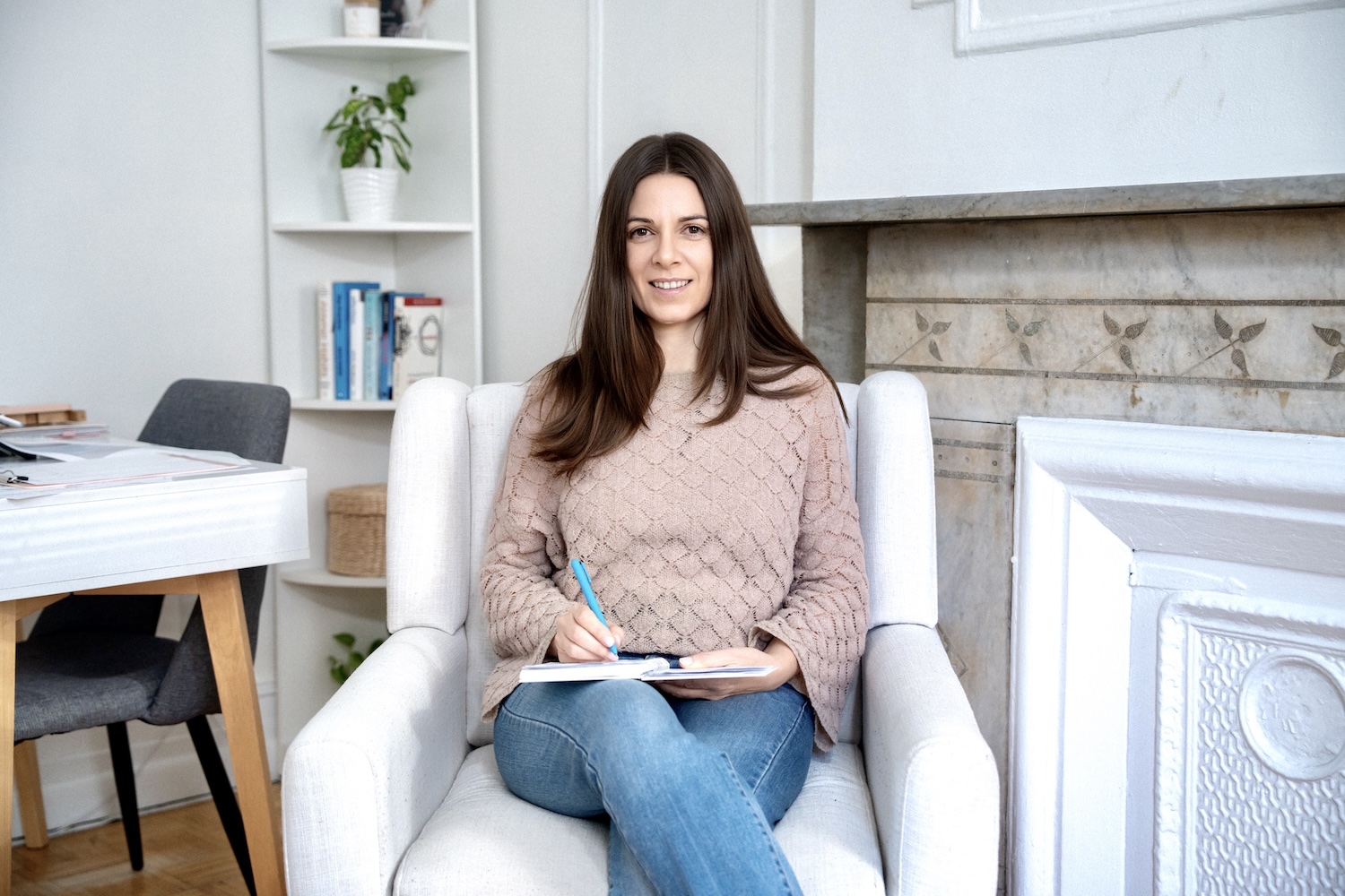 Woman with long brown hair sitting on a white armchair, writing in a notebook with a blue pen.