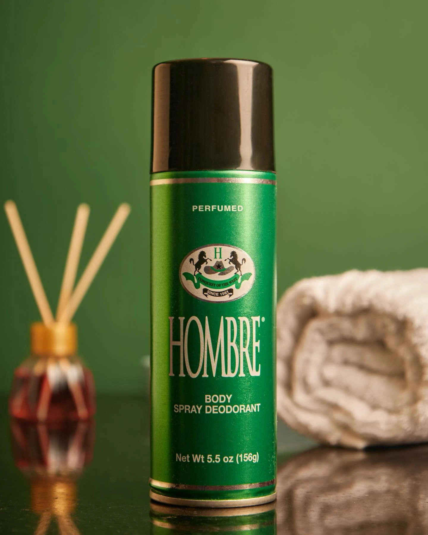 HOMBRE® Green deodorant body spray displayed with reed diffuser and towel — fresh masculine fragrance