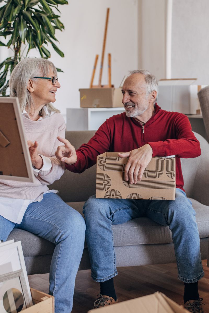 Smiling elderly couple sitting on a couch surrounded by moving boxes, the man holds a patterned box while the woman holds a framed picture.