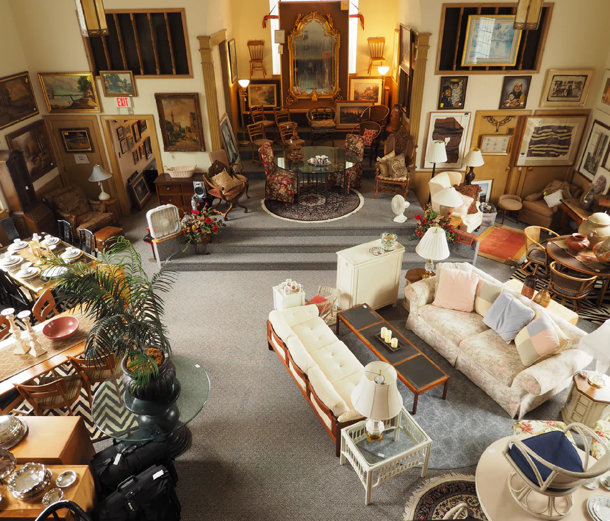 Antique furniture showroom with sofas, chairs, tables, lamps, framed paintings, and decorative items arranged in a spacious room.