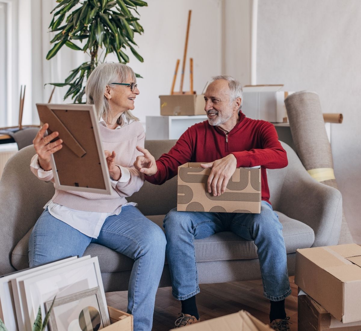 Smiling elderly couple sitting on a couch unpacking boxes and holding a picture frame in a room with moving boxes and decor.