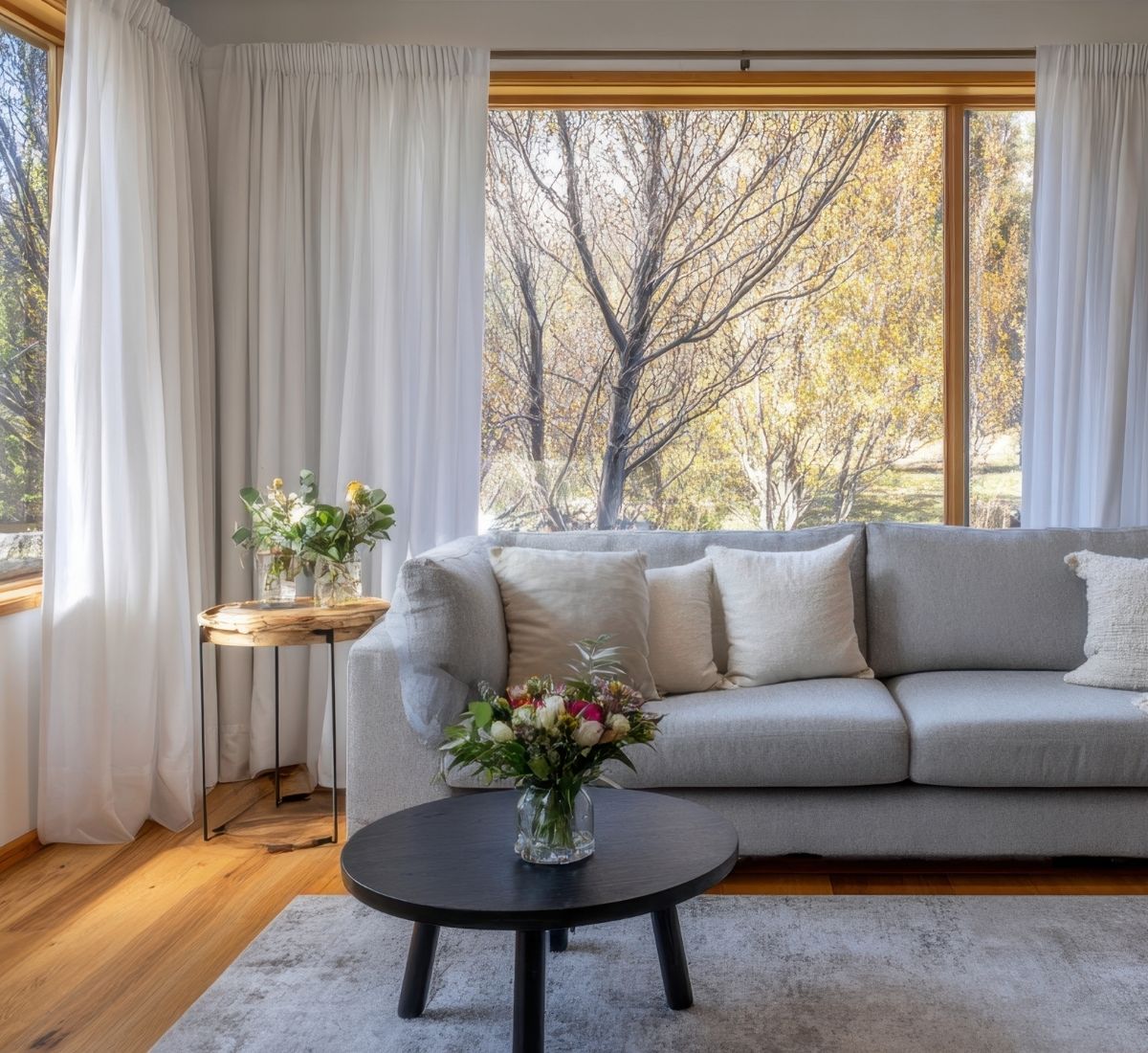 Living room with a light gray sofa adorned with cream pillows, a black round coffee table with a floral arrangement, wooden side table with flowers, and large windows showing autumn trees outside.