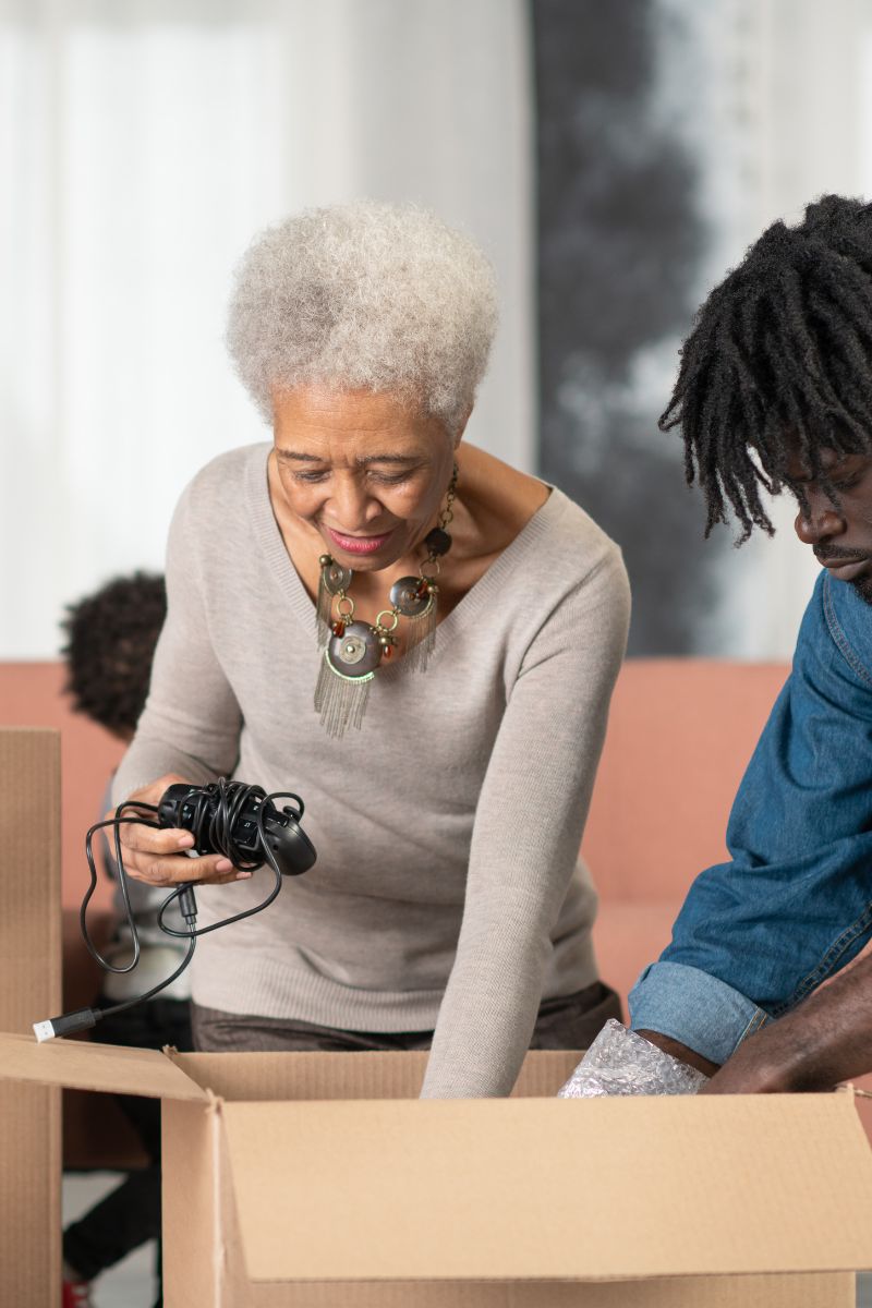 Elderly woman unpacking a cardboard box while holding a tangled game controller cord, with a man beside her looking into the box.