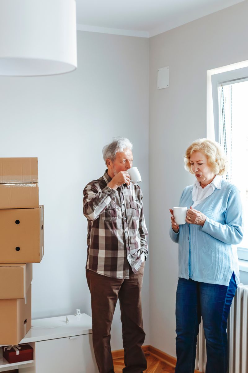 Older man and woman standing in a room with moving boxes, each holding a white mug and drinking coffee.
