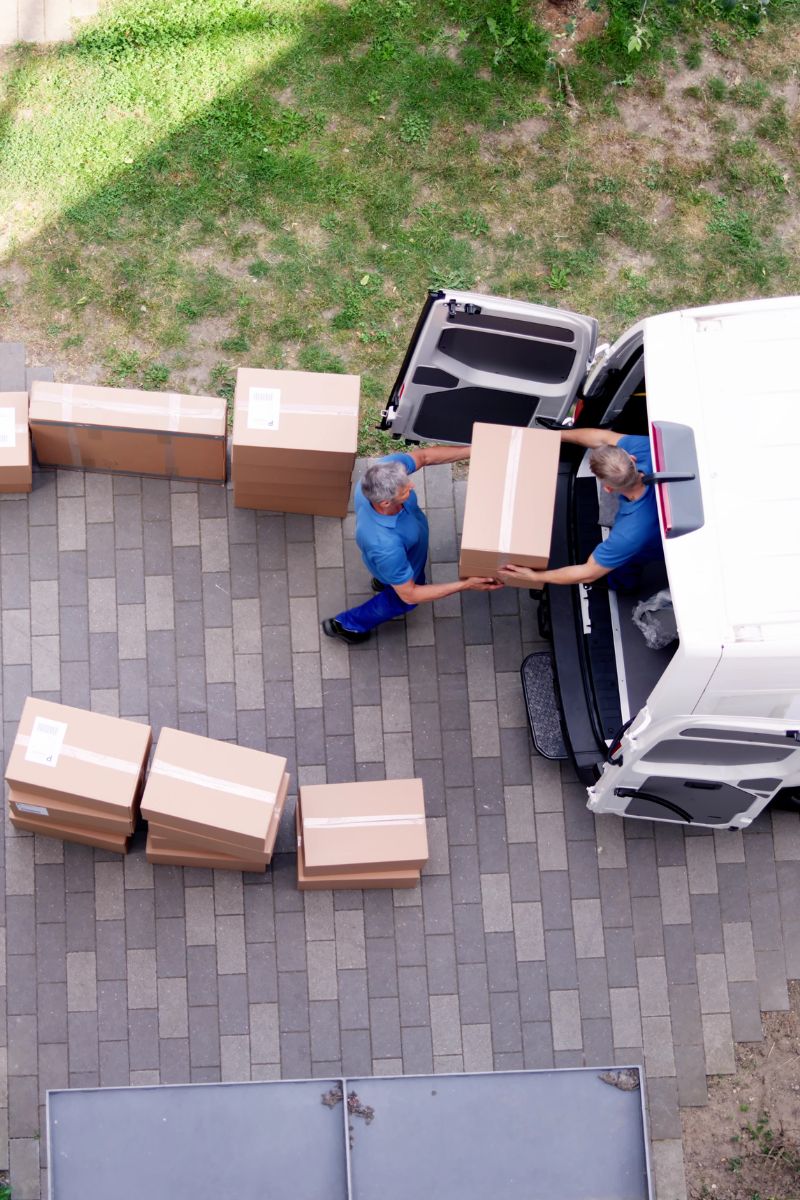 Two men in blue shirts unloading and passing cardboard boxes from a white delivery van on a paved driveway.