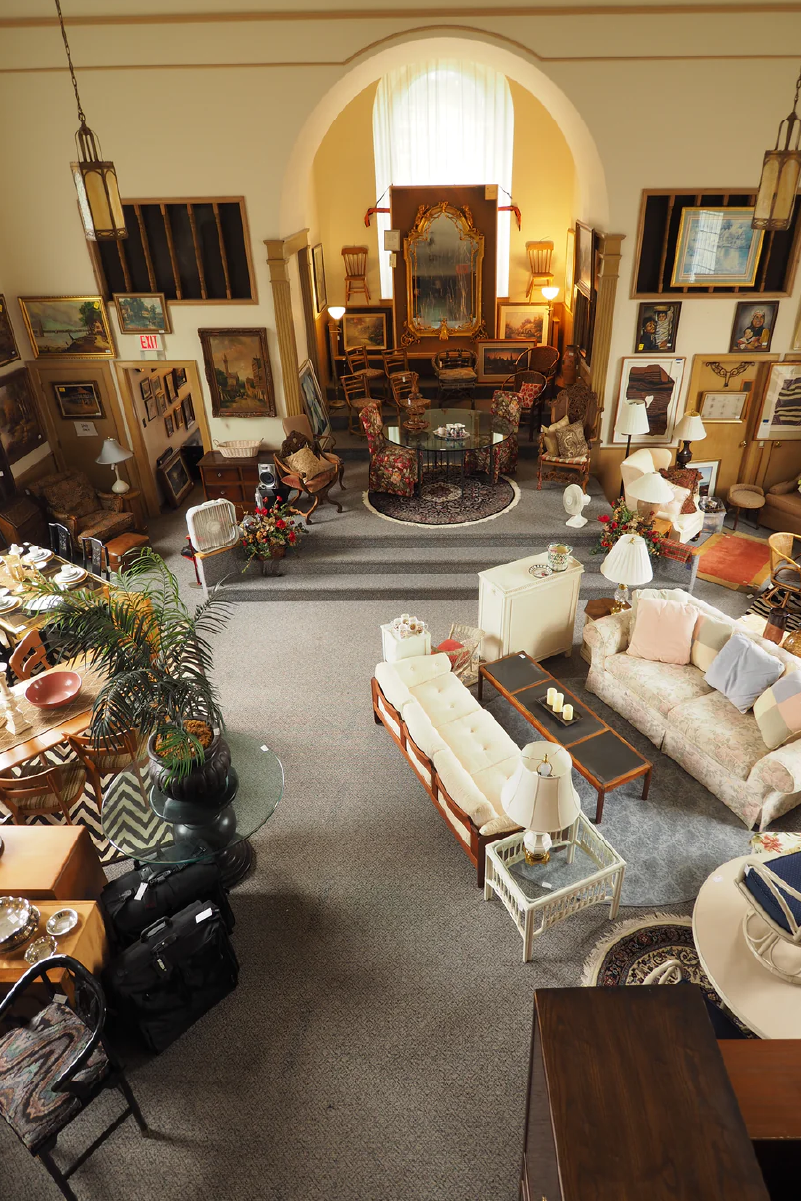 View of a vintage furniture store showroom with sofas, chairs, tables, framed paintings, and decorative lamps.