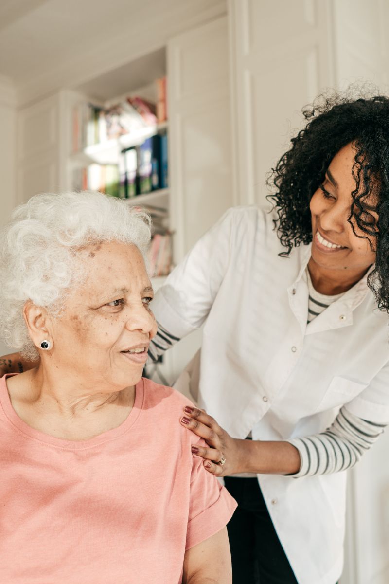 Smiling female caregiver in white coat offers support to elderly woman with white curly hair wearing a pink shirt.