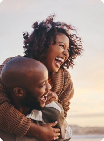 Un couple joyeux riant ensemble, la femme enlacée derrière l'homme avec un paysage au coucher du soleil en arrière-plan.