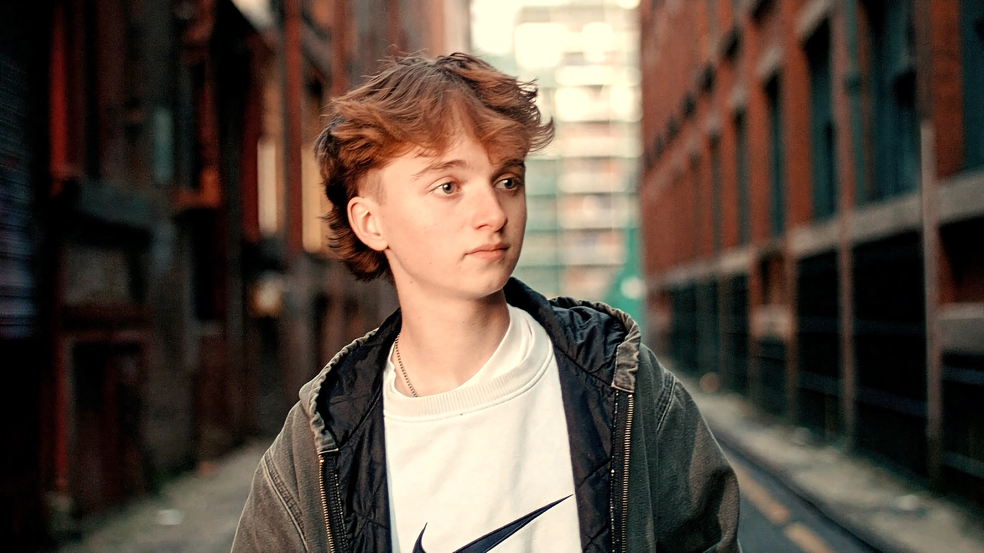 Young person with short brown hair wearing a white Nike sweatshirt and dark jacket in an urban alleyway.