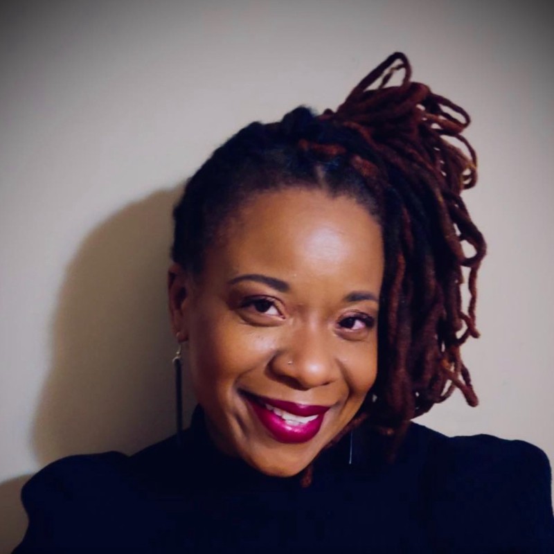 Portrait of a smiling woman with dark skin and long reddish-brown dreadlocks styled to one side, wearing dark lipstick, a nose ring, long earrings, and a black top against a plain light background.