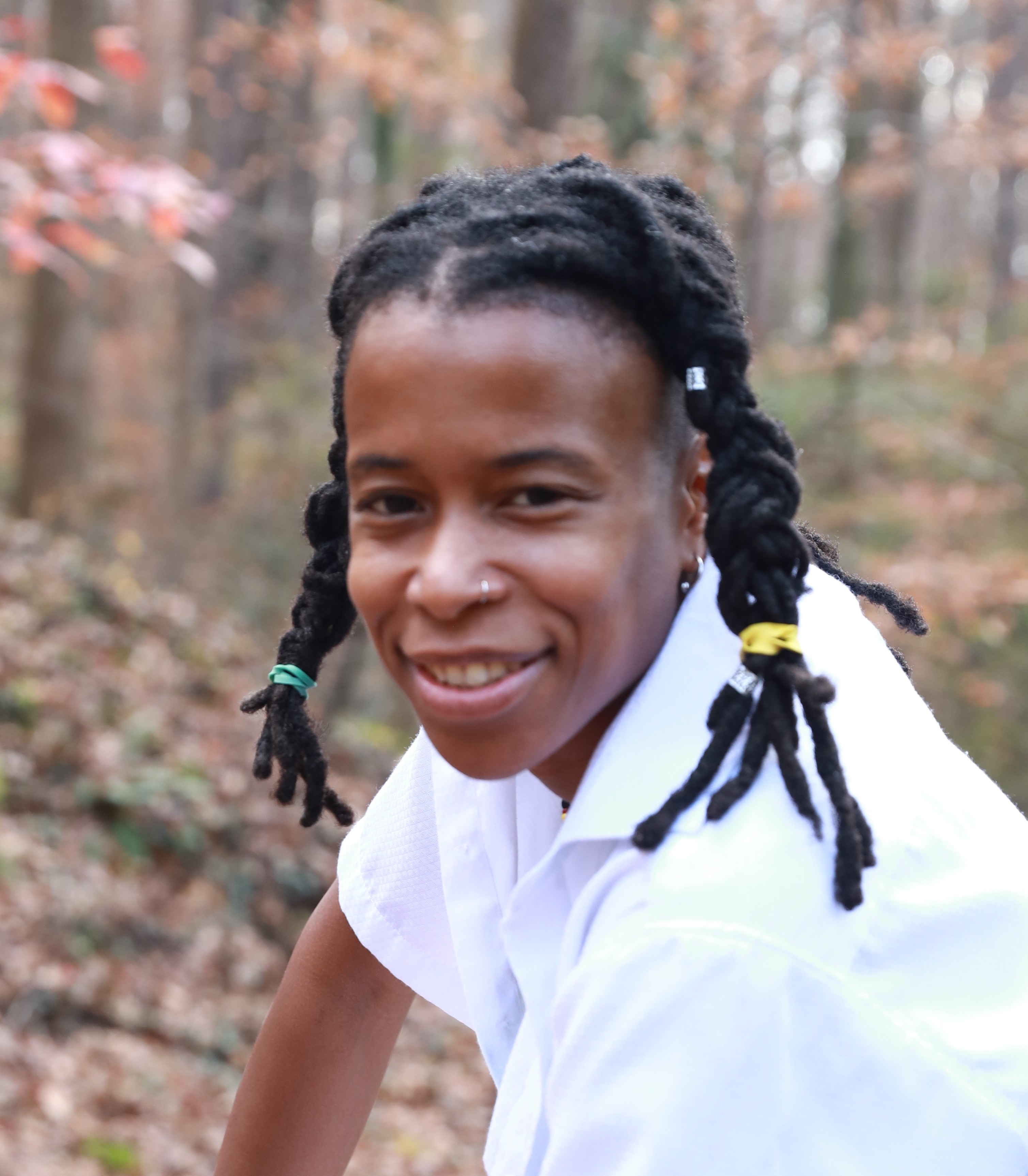 Smiling person with dark braided hair tied with colorful bands, wearing a white shirt, outdoors in a wooded area with brown and green foliage.