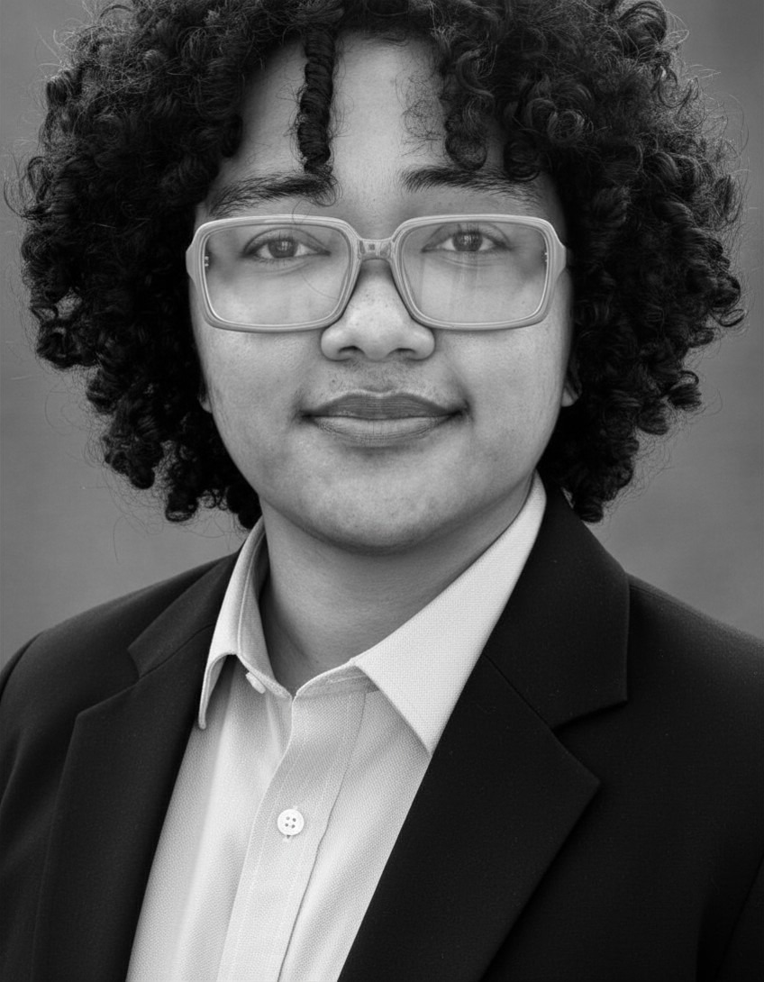 Black and white headshot of a person with curly hair wearing large eyeglasses, a light-colored collared shirt, and a dark blazer, looking directly at the camera with a neutral expression.