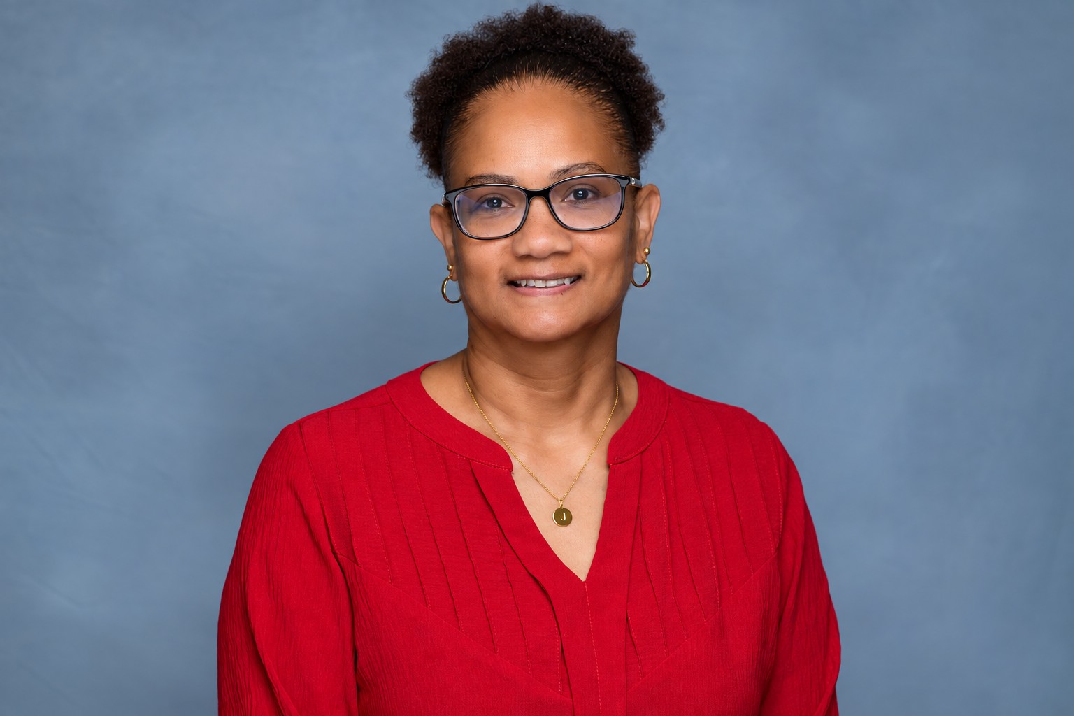 Smiling woman with short curly hair wearing glasses, gold hoop earrings, a red top, and a gold necklace with a circular pendant.