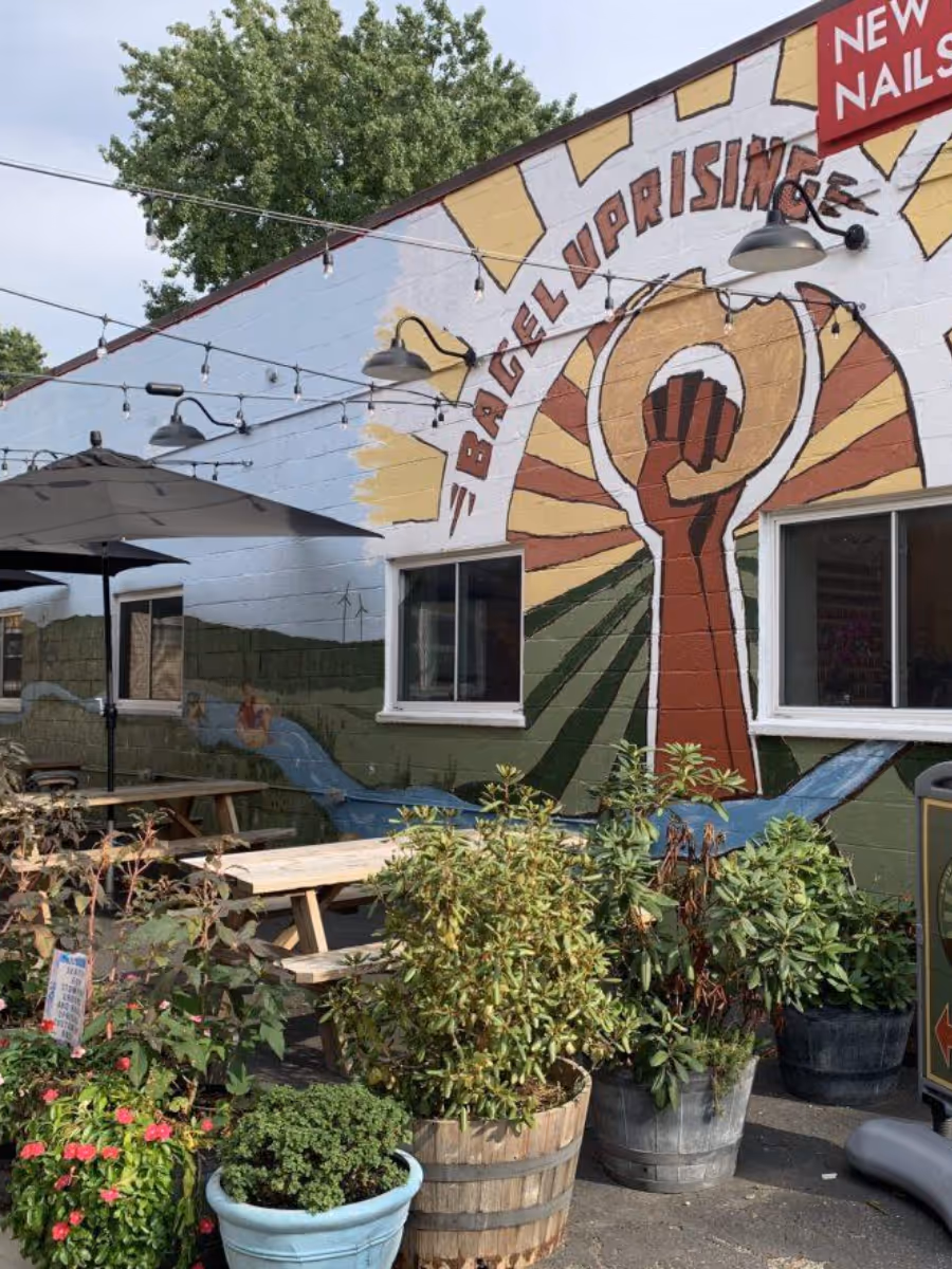 Outdoor seating area with wooden picnic tables, potted plants, and a mural of a raised fist and the words 'Bagel Uprising' on the building wall.