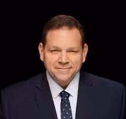 Portrait d'un homme souriant en costume bleu marine et cravate à pois sur fond noir.