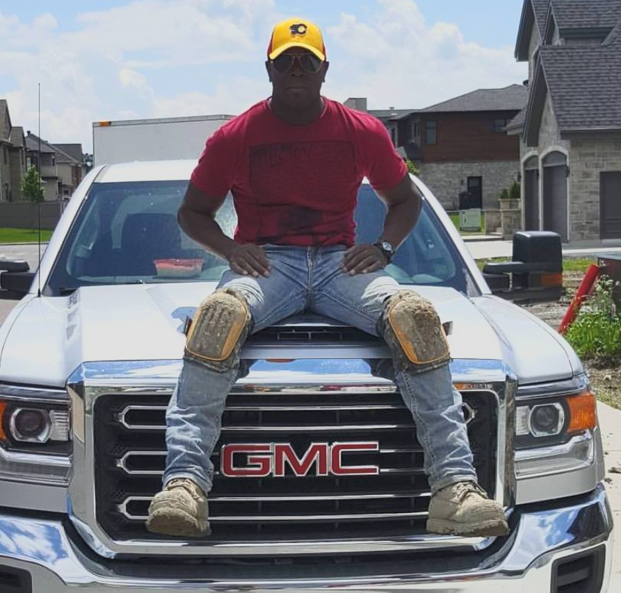 Un homme portant une casquette jaune, un t-shirt rouge, des genouillères et des bottes de travail est assis sur le capot d’un grand camion blanc GMC dans un quartier résidentiel.