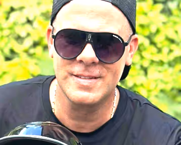 Homme portant des lunettes de soleil noires et un t-shirt noir avec un fond flou de feuillage vert.