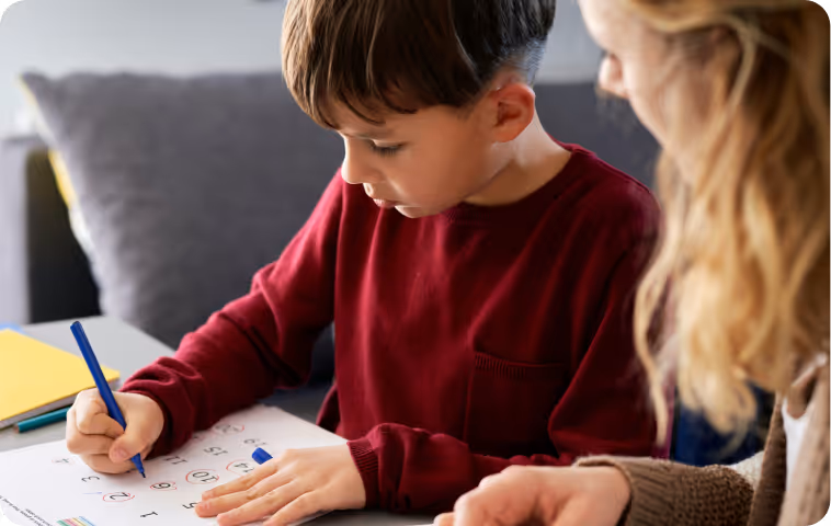 Young boy in red sweater focusing on his homework while a woman beside him offers support.