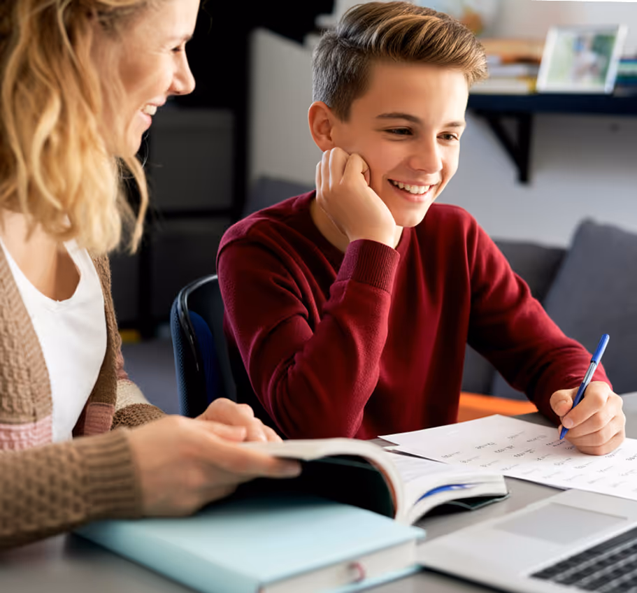 Teenage boy smiling while writing on paper at a desk with a woman holding a book beside him.
