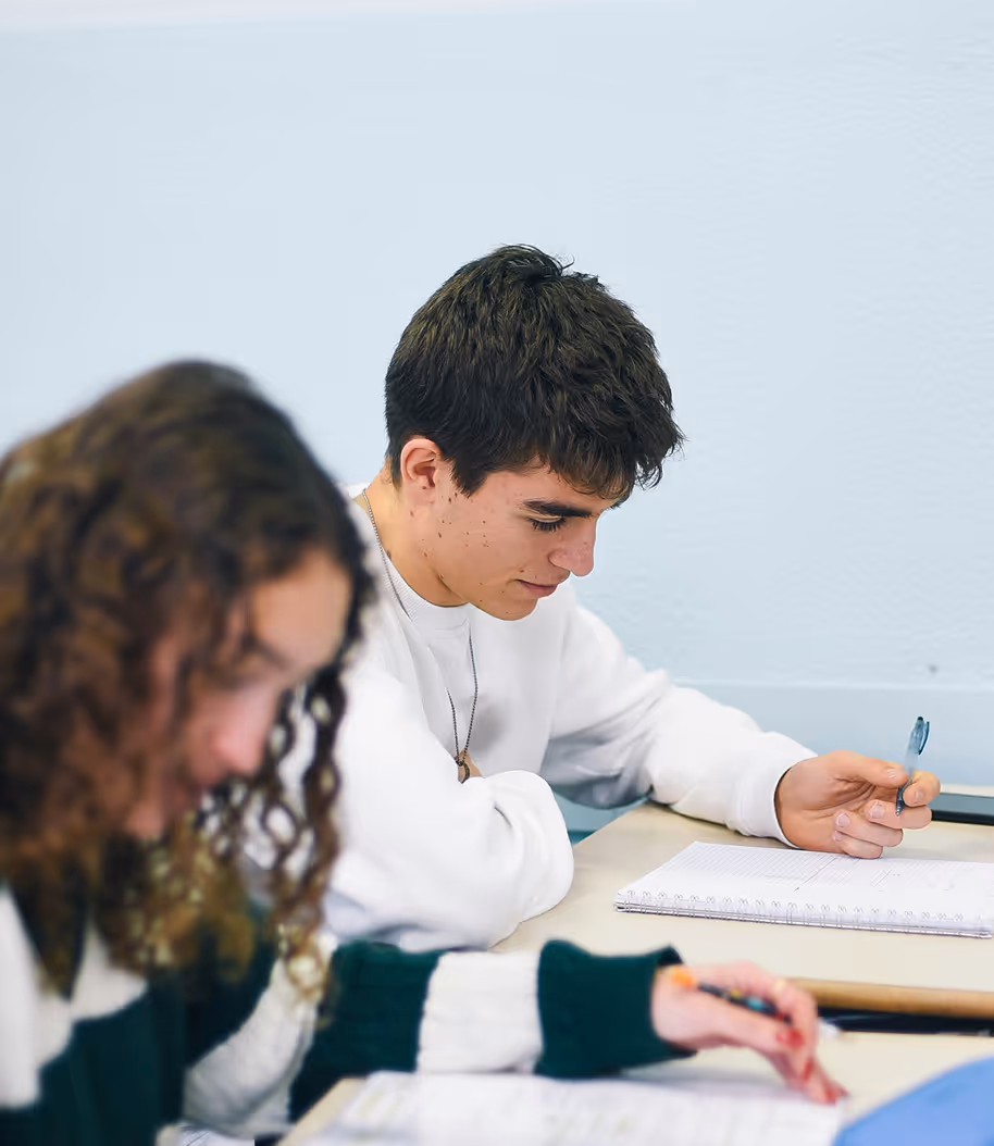 Two students sitting at desks, focused on writing in notebooks during class.