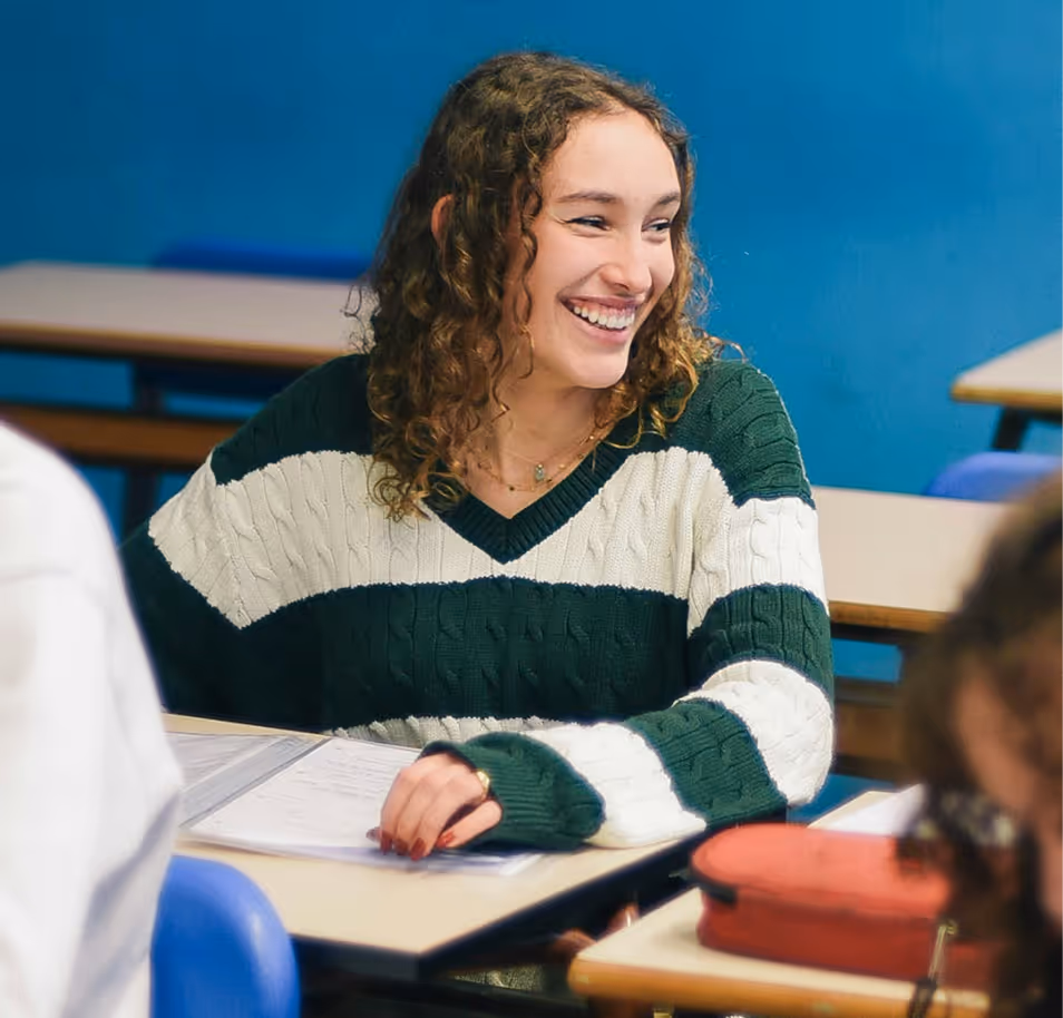 Smiling young woman with curly hair wearing a green and white striped sweater sitting at a classroom desk with papers.