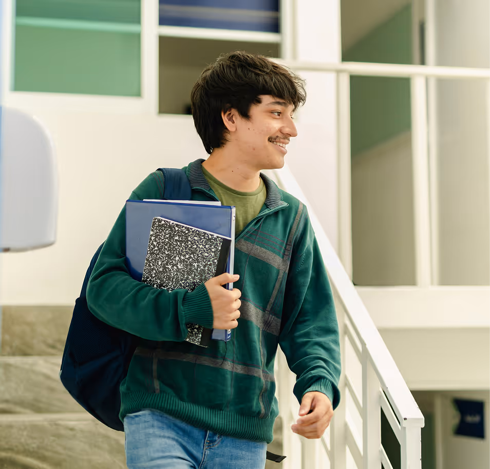 Smiling young man carrying notebooks and a backpack walking indoors near stairs and windows.