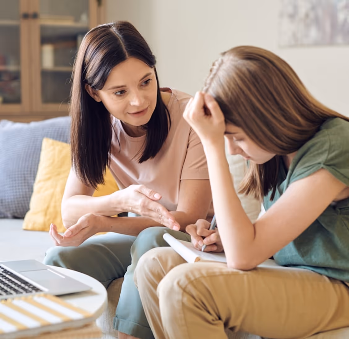 Woman calmly explaining something to a distressed teenage girl who is writing on a notebook.