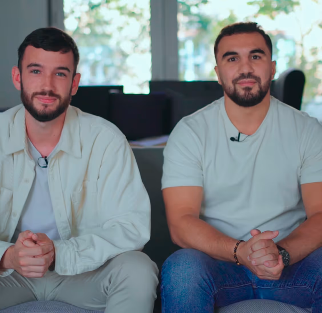 Two men sitting side by side on a couch indoors, both with clasped hands and looking at the camera.