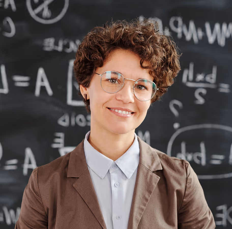 Smiling woman with short curly hair and glasses standing in front of a chalkboard with mathematical equations.