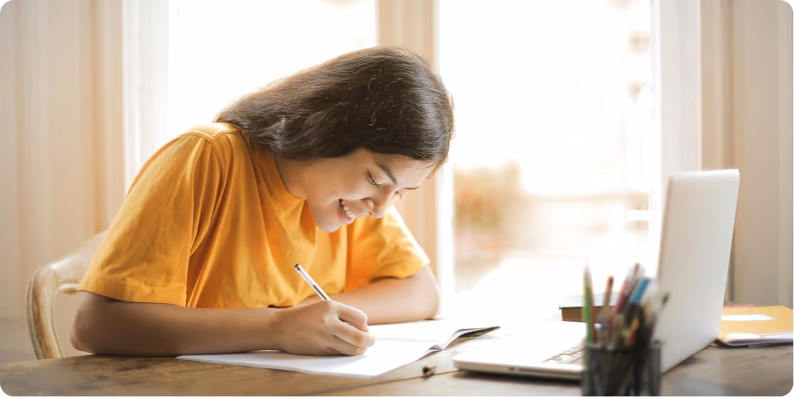 Smiling young woman in a yellow shirt writing in a notebook at a table with a laptop and pencil holder in a bright room.