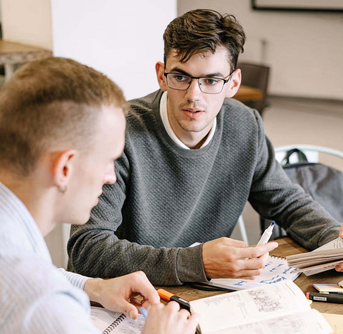 Two young men engaged in a discussion over notebooks and papers at a table.