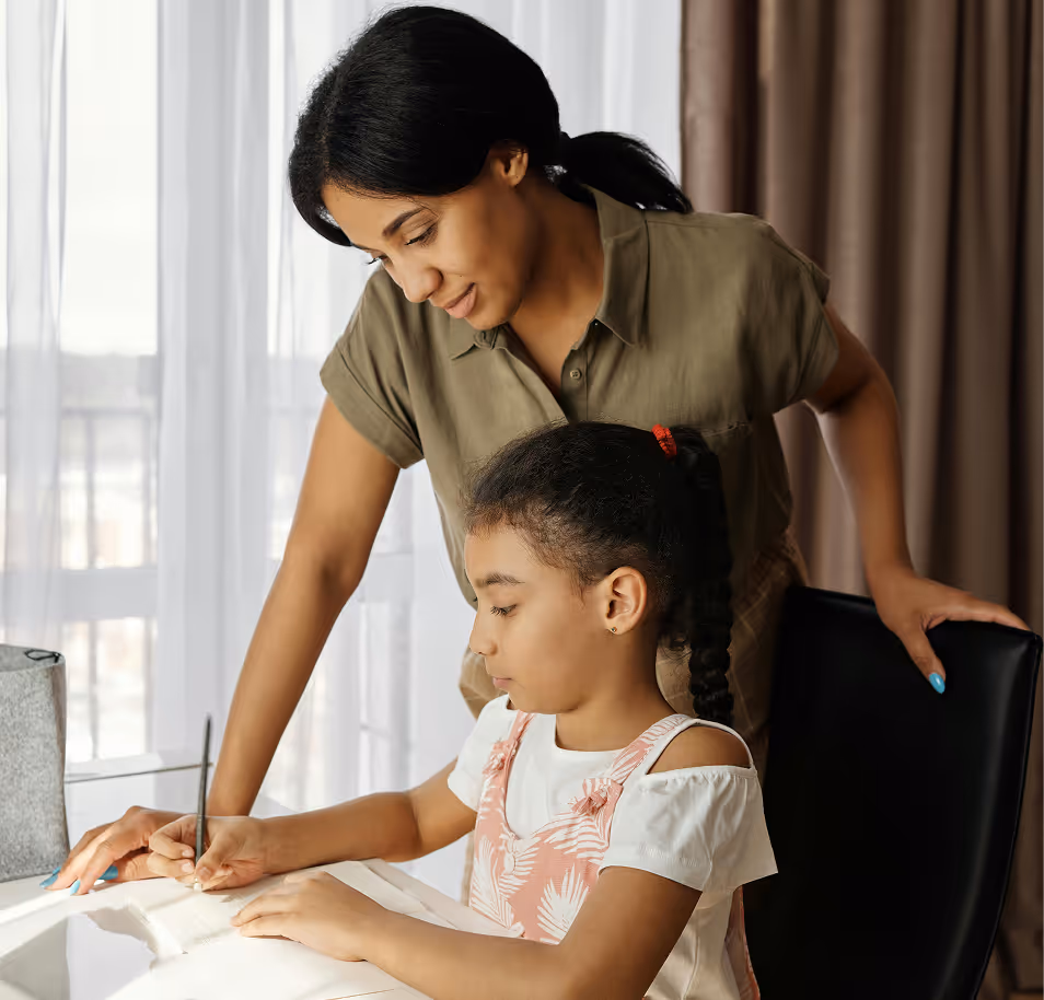 Mother attentively helps young daughter with writing at a table near a window with curtains.
