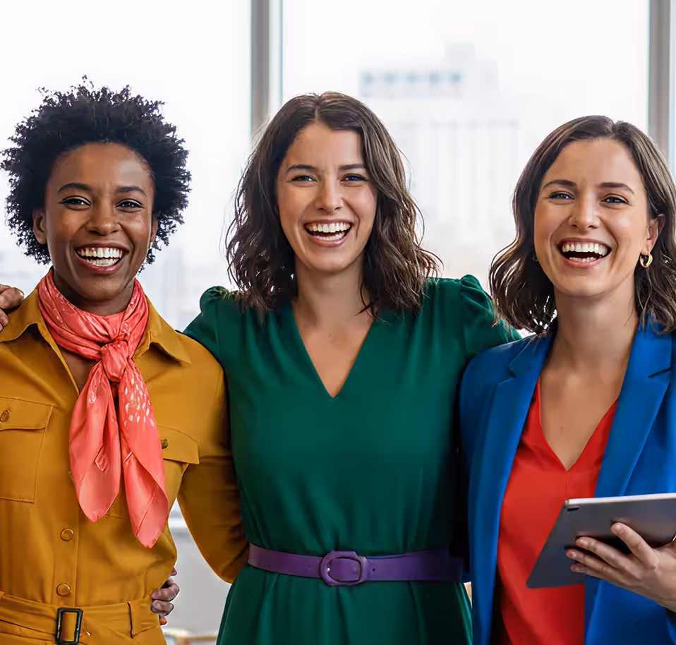 Three smiling women standing close together in colorful professional attire in a bright office.