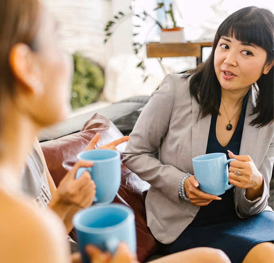 Three women sitting and talking on a couch, each holding a light blue coffee mug.