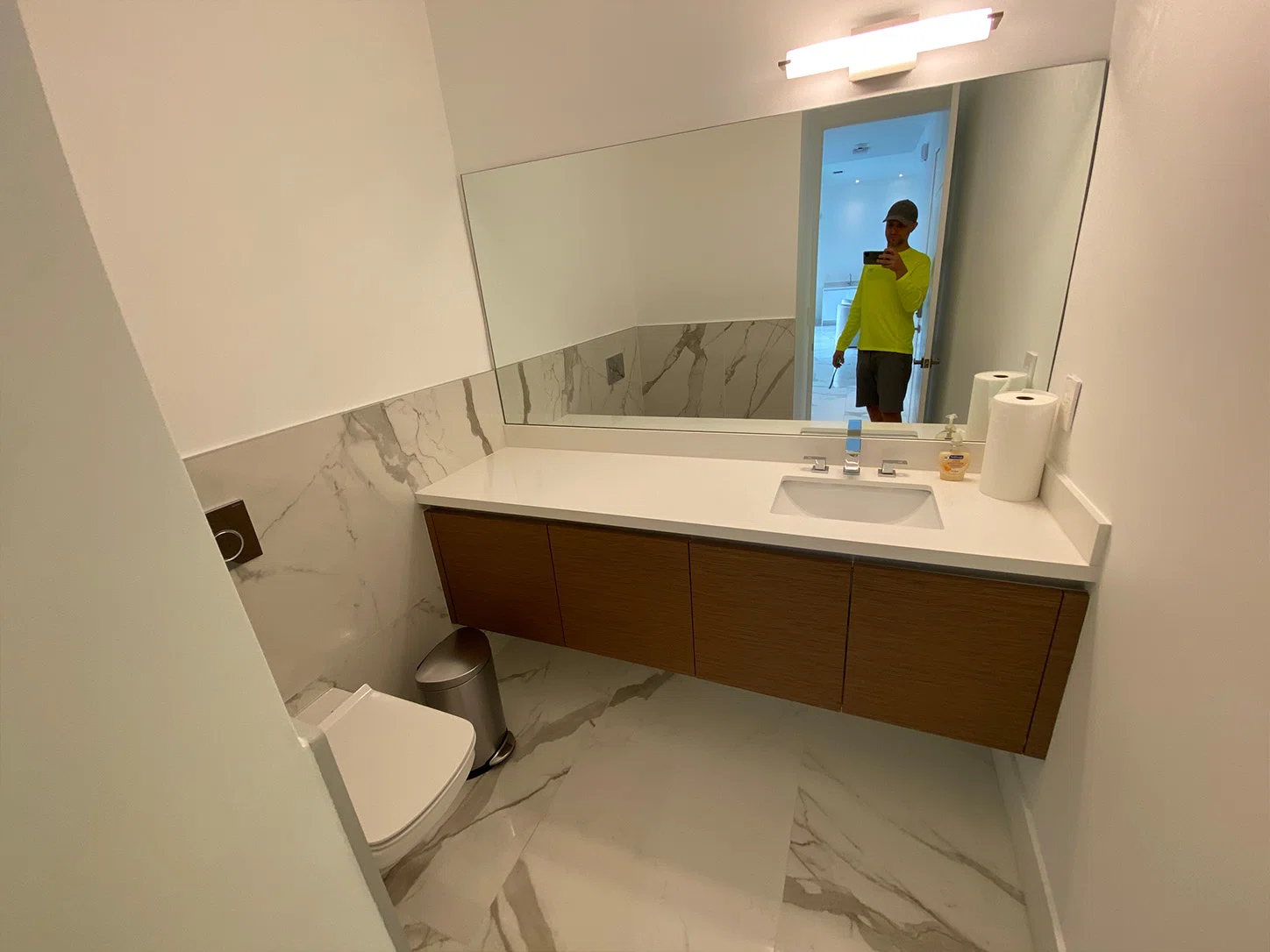 Powder room before remodeling in Weston, FL, with a floating wood vanity, white countertop, large rectangular mirror, and marble-look tile.