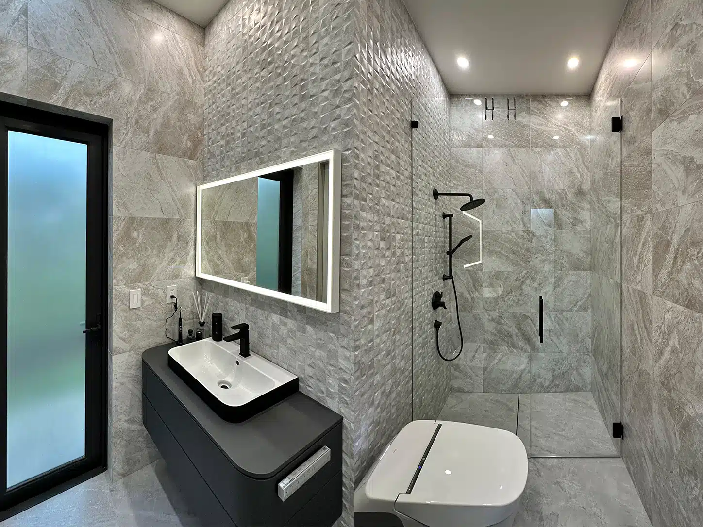 Remodeled bathroom in Weston, FL, with textured gray tile, a floating charcoal vanity, LED mirror, and glass shower with black fixtures.