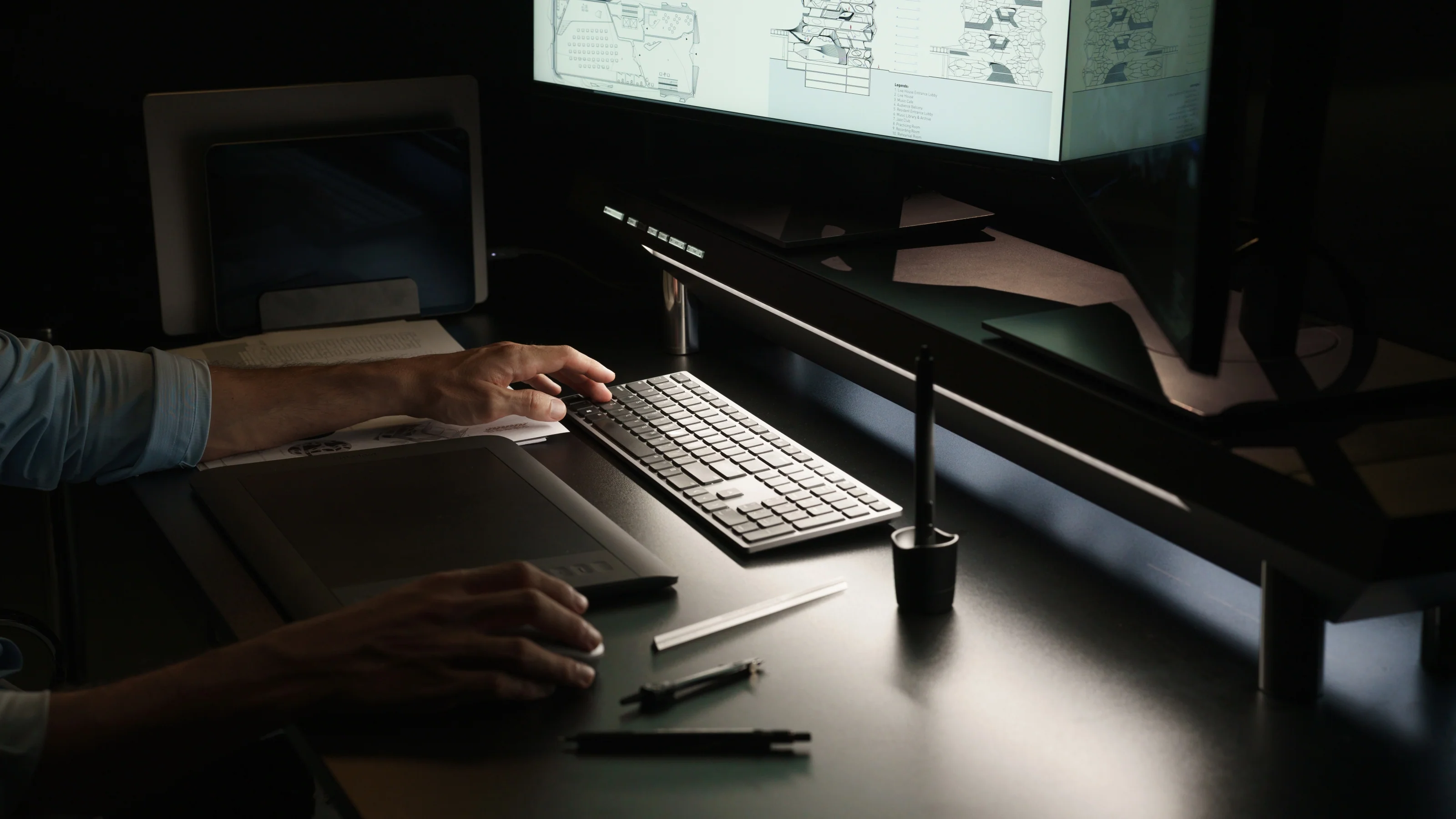 Person using a keyboard and mouse on a desk with a drawing tablet, stylus, and dual monitors displaying architectural plans.
