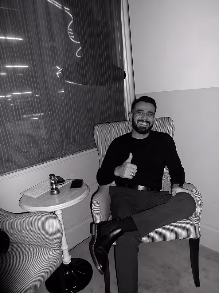 Cristian Maldonado smiling and giving thumbs up while sitting cross-legged in a striped armchair next to a small round table with a lantern and phone.