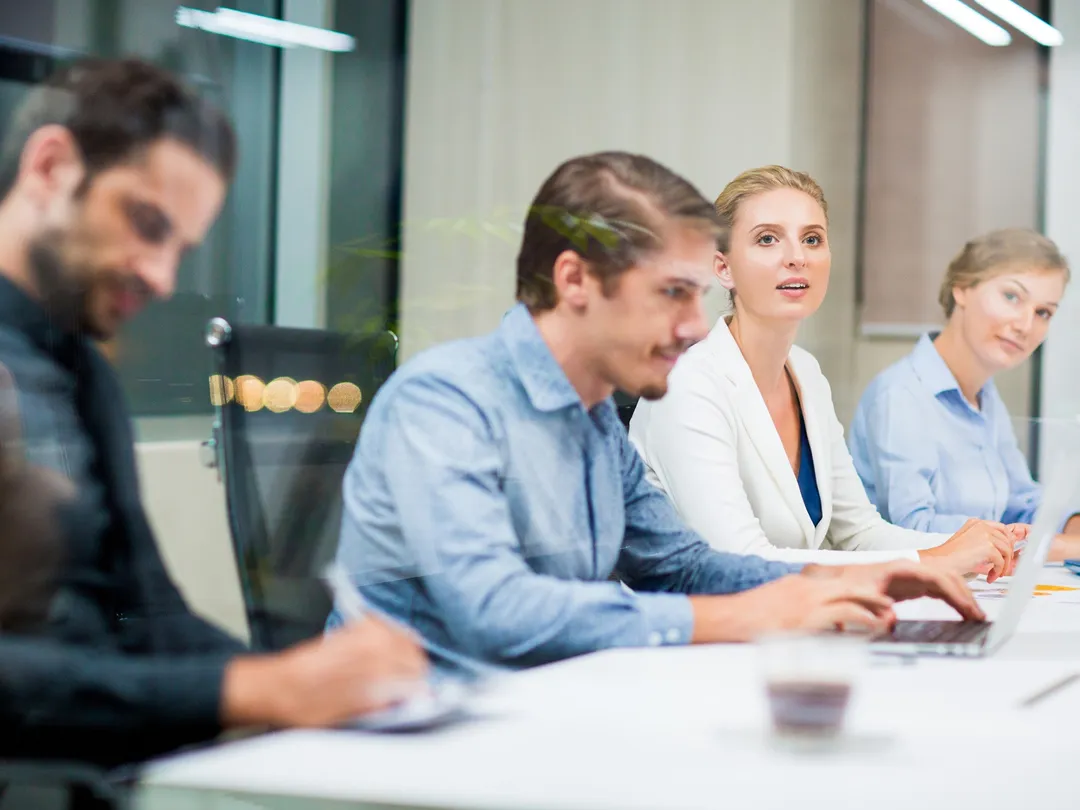 Four business professionals engaged in a meeting, with two men and two women seated at a table using laptops and taking notes.