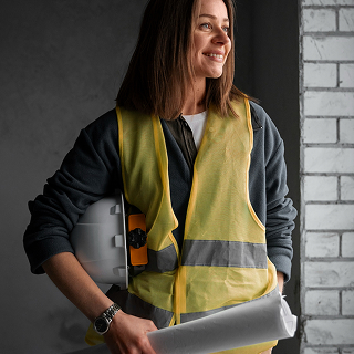 Smiling woman in a yellow safety vest holding a white hard hat and a rolled-up blueprint, standing by a brick wall.