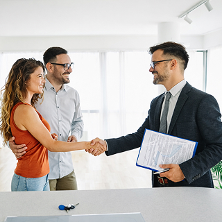 Real estate agent shaking hands with a smiling couple in a bright empty apartment, with keys and laptop on the counter.