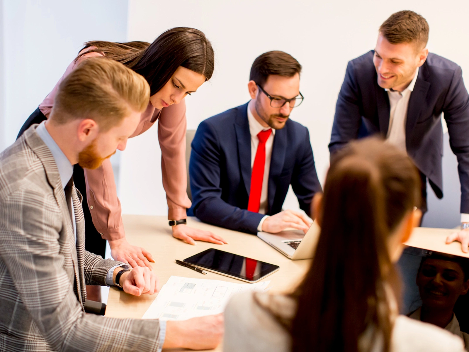A group of five business professionals collaborating around a table with documents, a laptop, and a tablet.