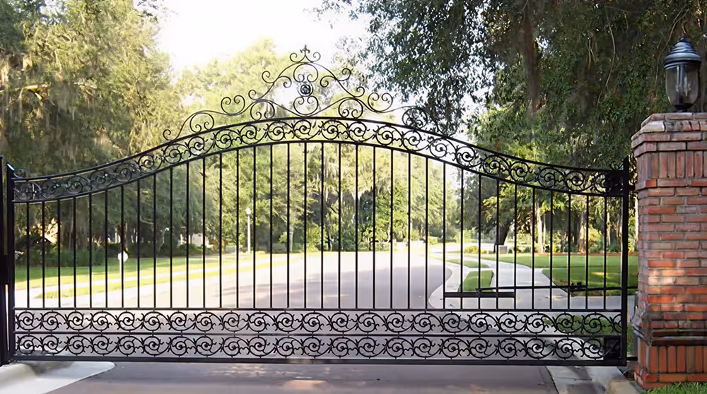 Ornate black wrought iron gate with scrollwork and brick pillar, blocking a tree-lined road.