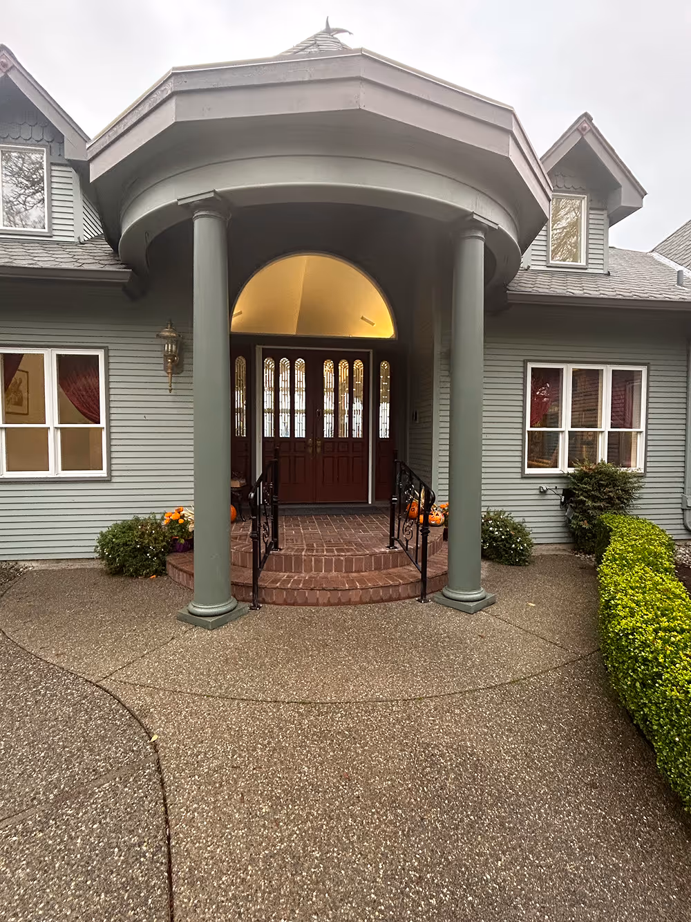 Front entrance of a gray house with two large round columns, brick steps, and double wooden doors with glass panels under an arched ceiling.
