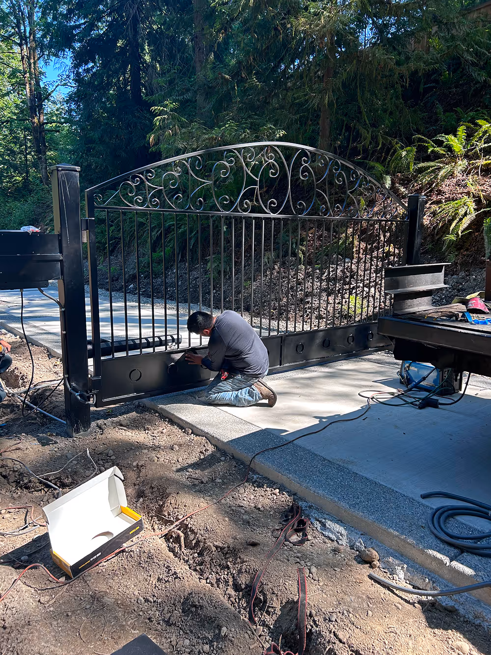 Man kneeling and working on installing a large black decorative metal gate on a concrete driveway in a wooded area.