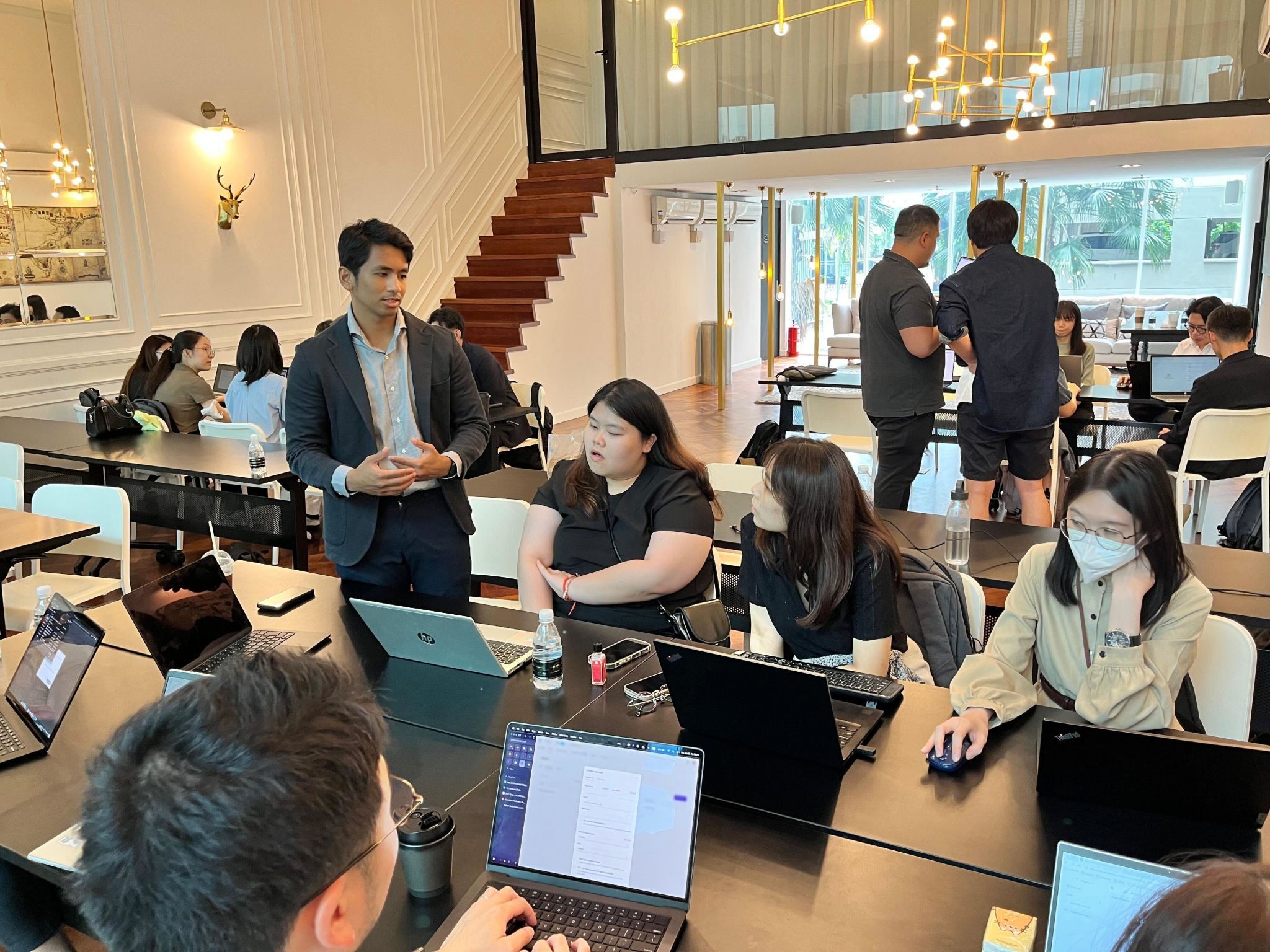 Group of people working on laptops and discussing in a modern office with large windows and wooden stairs.