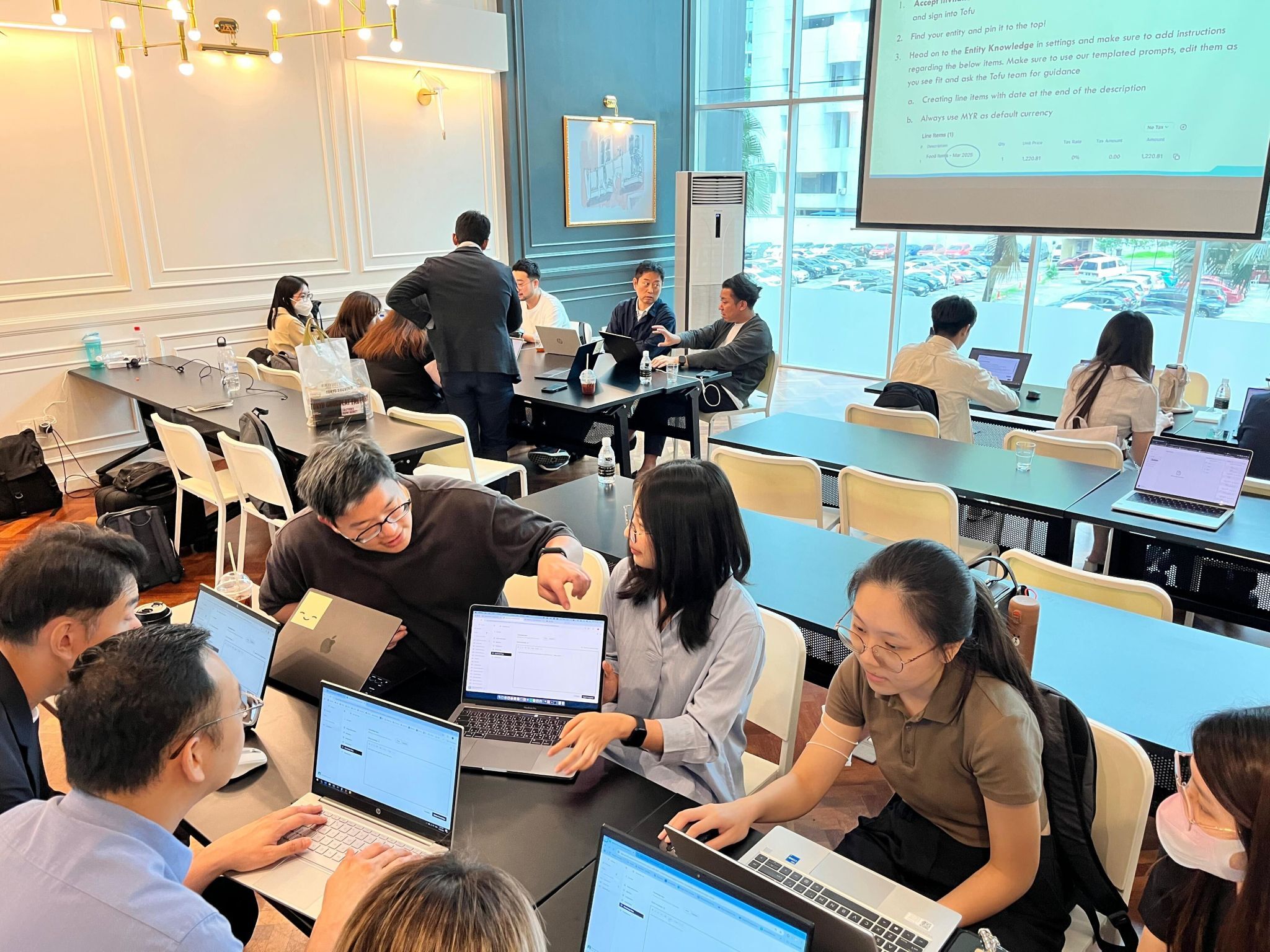 Group of people working collaboratively on laptops in a modern conference room with a large window and projection screen.